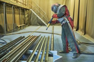 Industrial cleaning with vapor abrasive blasting equipment. The image shows a worker using a vapor abrasive blasting machine to clean a large metal surface, with protective gear and equipment in a workshop environment.