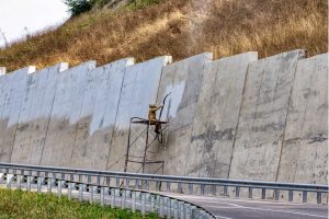 Vapor abrasive blasting for road wall cleanup. The image shows a worker using a vapor abrasive blasting machine to clean a road wall, with dust and debris being removed from the surface in an outdoor setting.